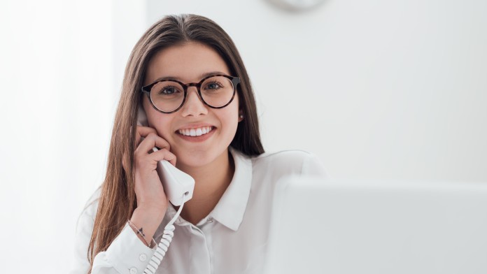 Lächelnde junge Frau am Telefon, sie sitzt an einem Schreibtisch und arbeitet mit einem Laptop 