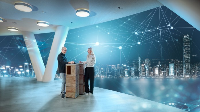 Zwei Personen stehen in einer Arbeitssituation an einem Stehtisch. Im HintegrTwo people are standing at a high table in a working situation. In the background is a brightly lit skyline with an abstract digital network.