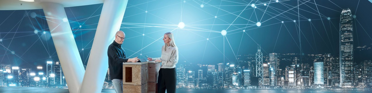 Two people are standing at a high table in a working situation. In the background, a brightly lit skyline with an abstract digital network