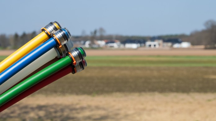 bunte Breitbandkabel draußen über Feld, Häuser verschwommen im Hintergrund