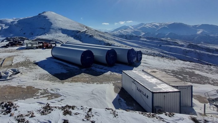 Tower parts of a wind turbine lie on the ground ready for assembly in a mountainous snowy landscape..