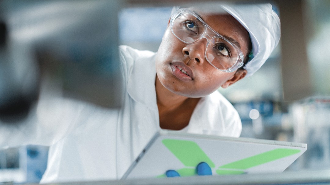 Eine Laborantin mit Schutzbrille analysiert offenbar Daten in einem Labor. In der Hand hält sie ein Tablet. Die Bildthemen sind Forschung, Qualitätssicherung und Innovation.